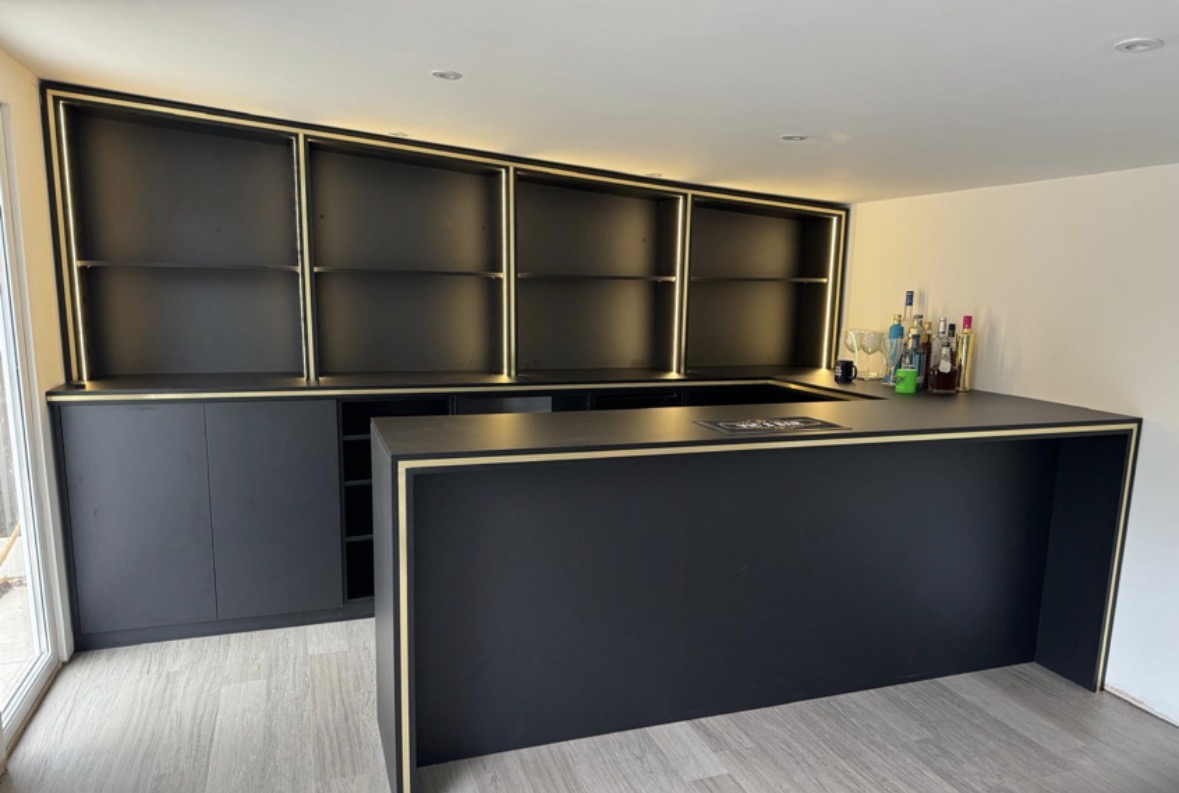 Graphite home bar with a brass-edged serving counter, wall of backlit display shelving shaped to the sloping ceiling and full-height cabinetry