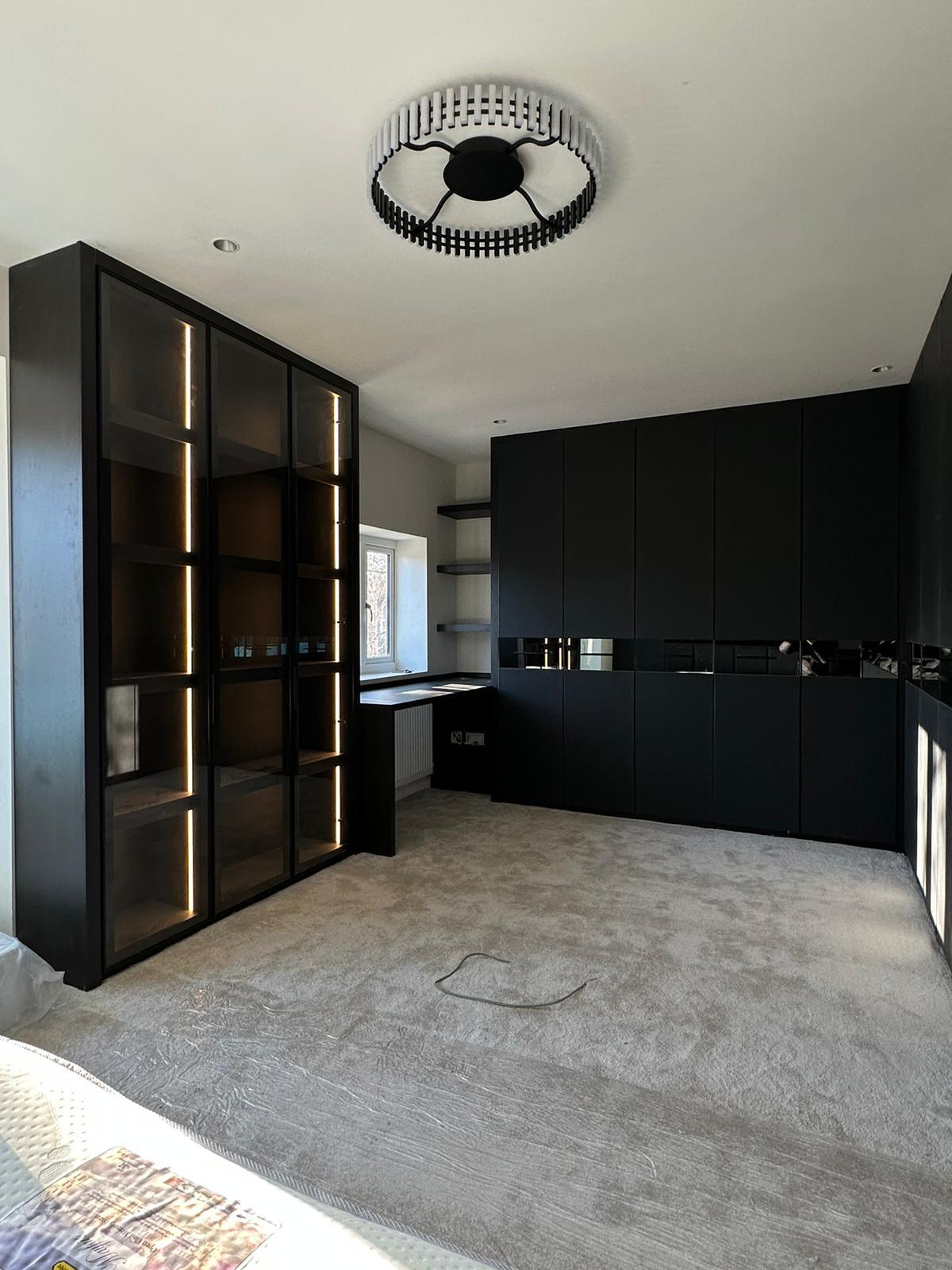 Matt black fitted bedroom furniture with a lit glazed display cabinet, mirrored door bands and a built-in desk under the window