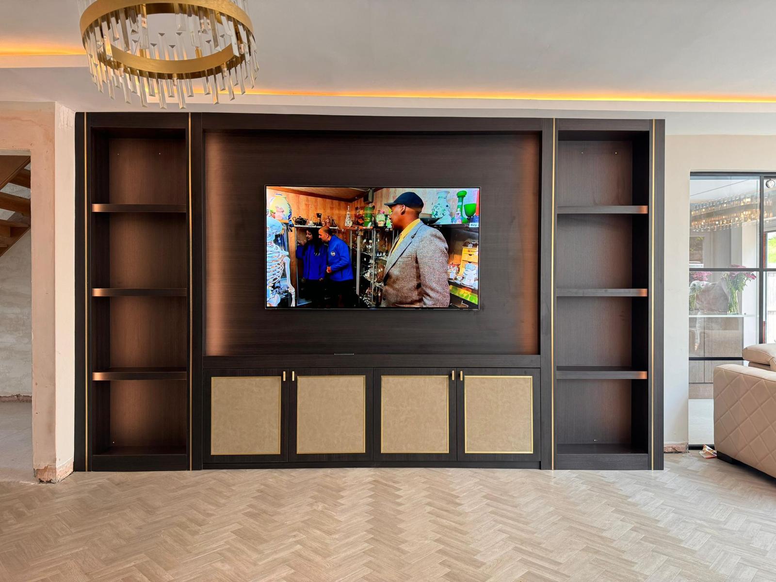Dark oak media wall with gold trim, open display shelving either side and suede-fronted cupboards beneath the television