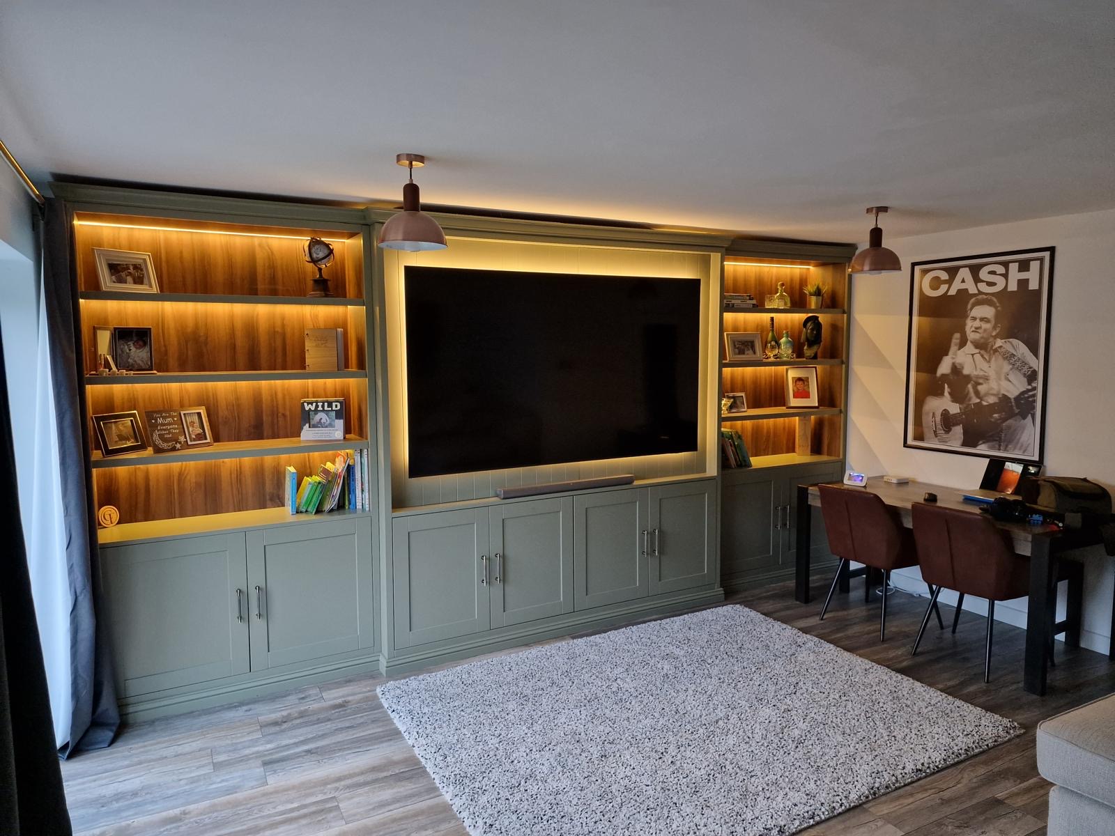 Sage green media wall with an inset television flanked by LED-lit walnut bookcases and framed artwork