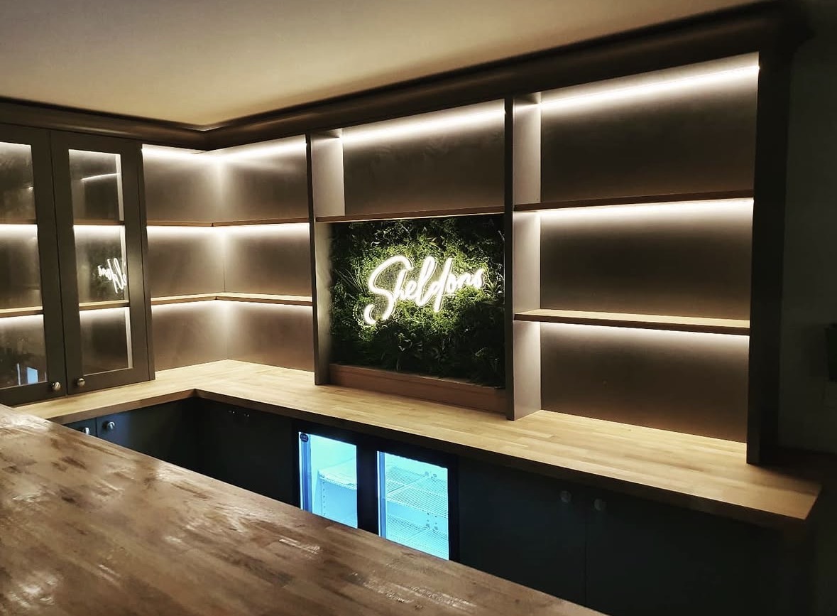 Backlit oak bottle shelving and glazed wall cabinets behind a home bar with integrated under-counter fridges