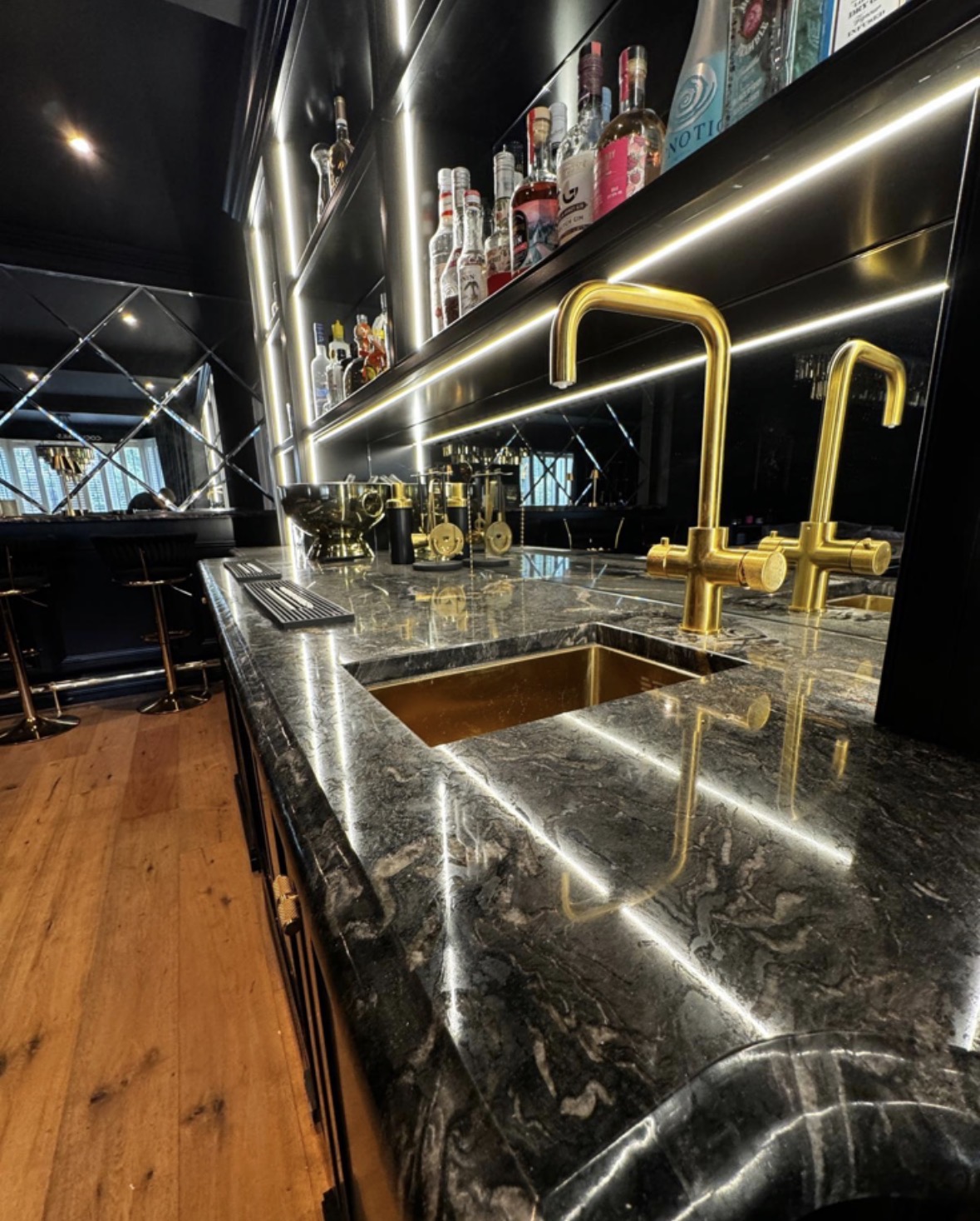 Detail of a green marble bar counter with a brushed gold tap and undermounted gold sink beneath LED-lit shelving