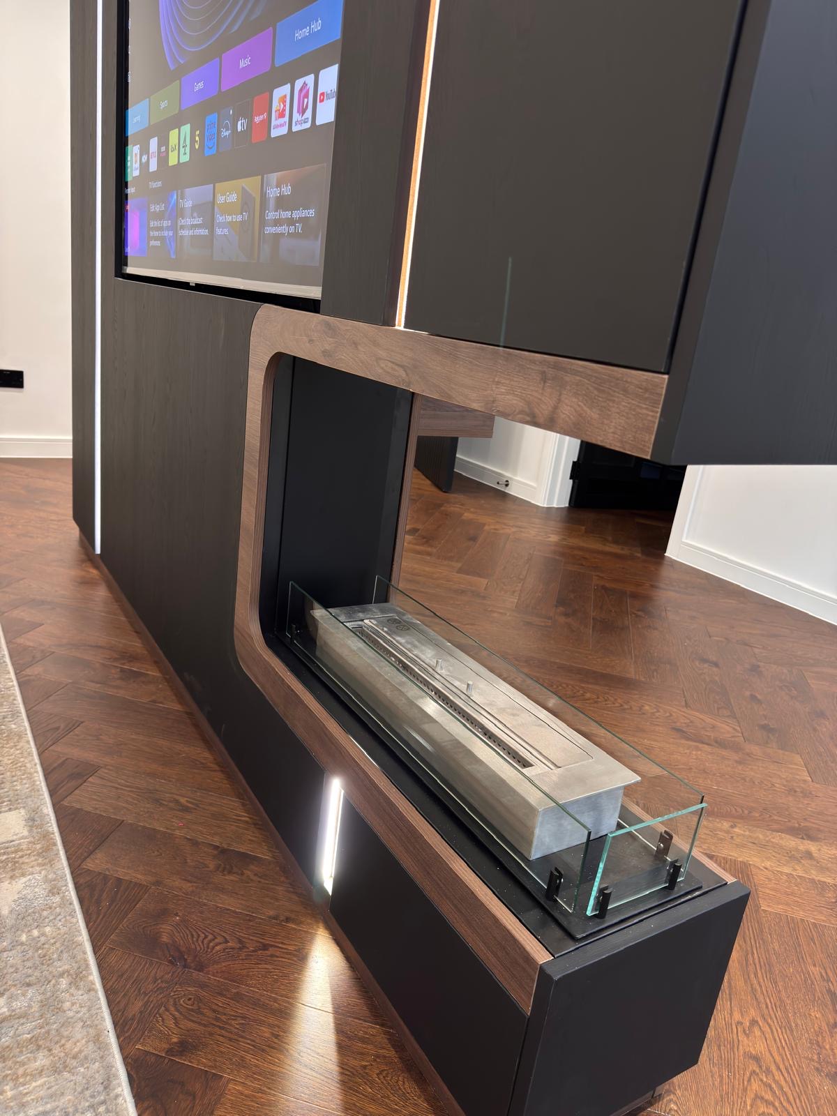 Close detail of a walnut-framed bio-ethanol fire with glass screen set into a matt black bespoke media wall, with the television above