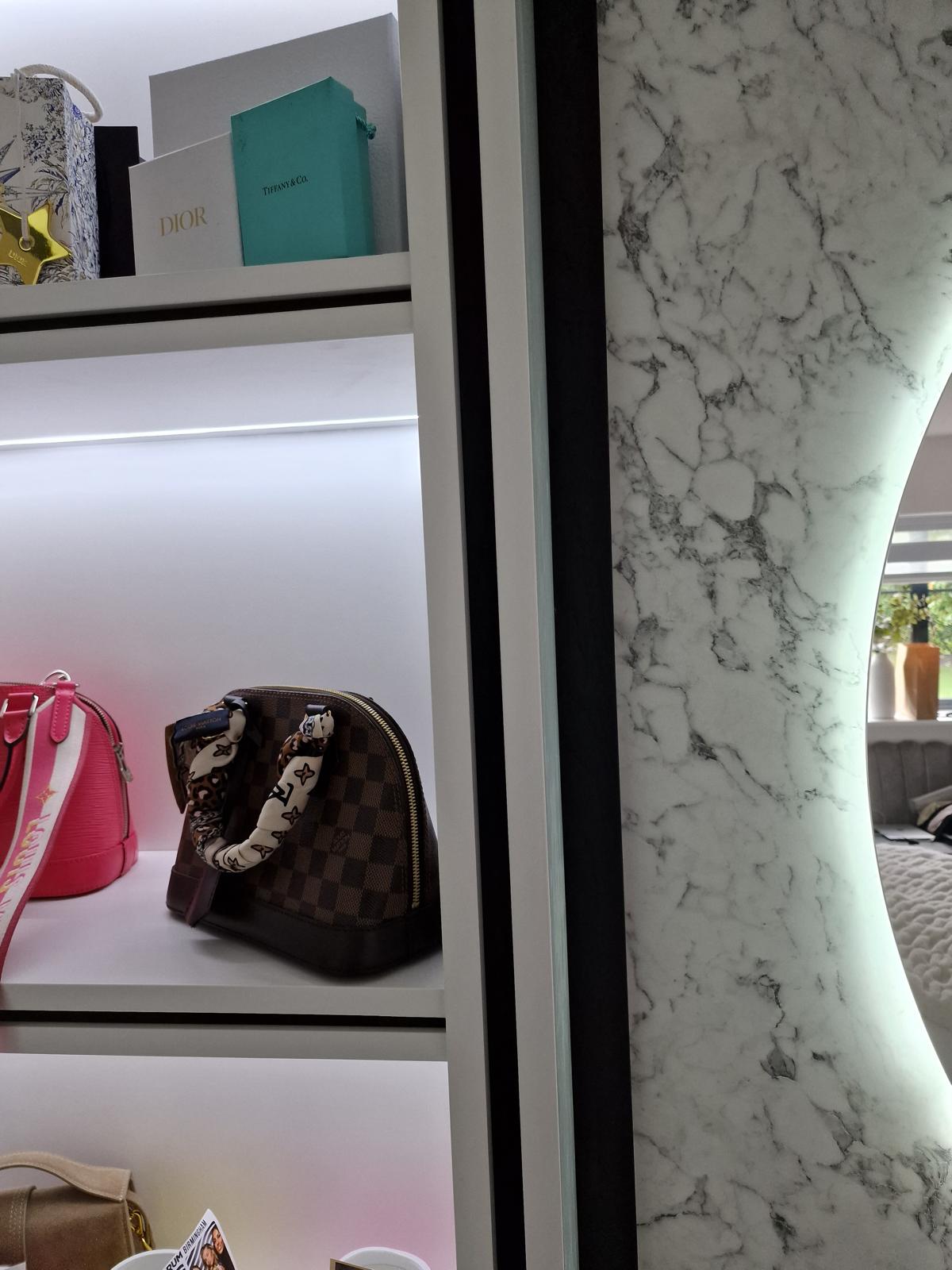 LED-lit glazed display shelving holding handbags and boxes beside a marble panel and backlit round mirror