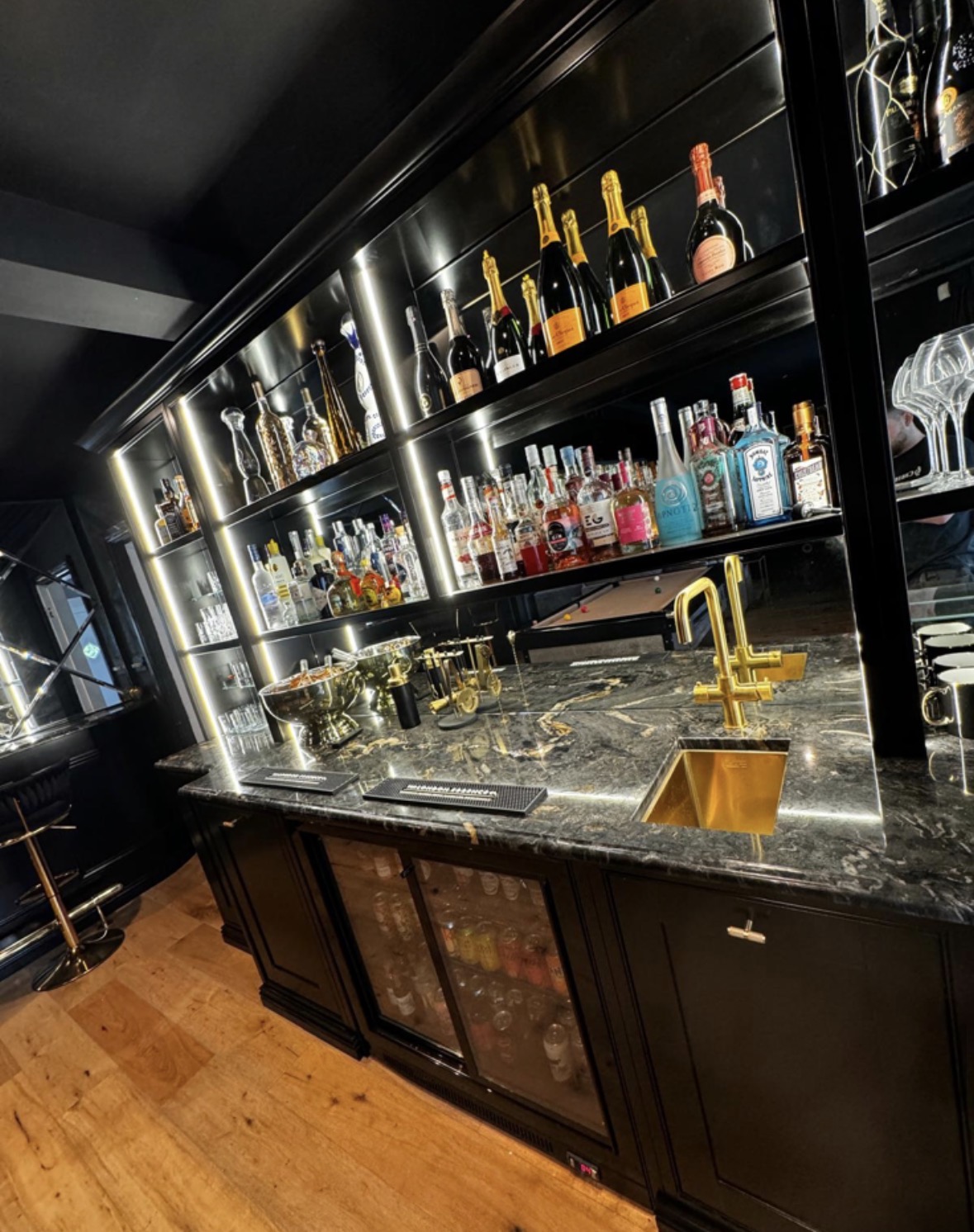 Curved black home bar with LED-lit bottle and glassware shelving, marble counter, brass tap and integrated glass-fronted bottle fridges