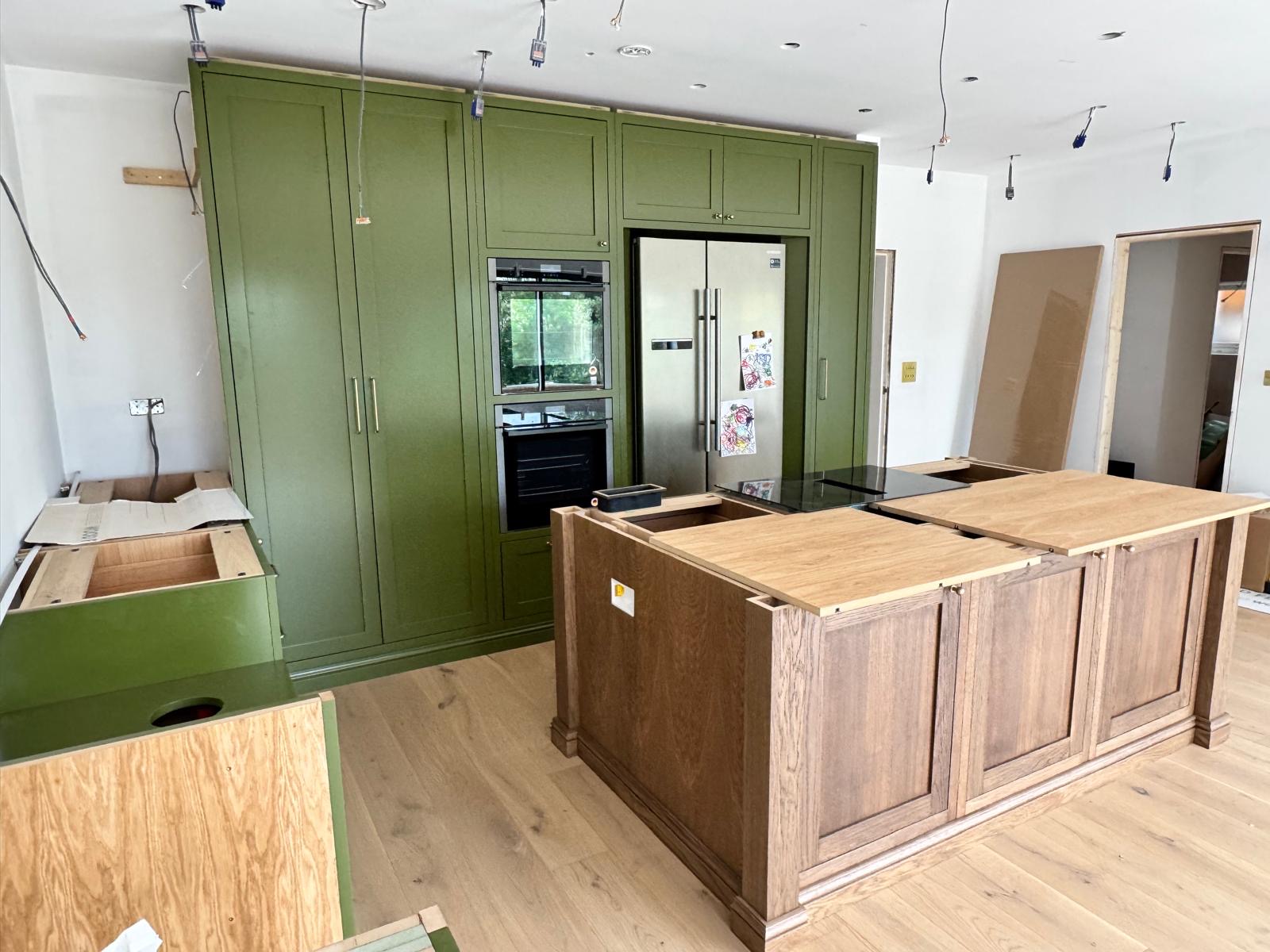 Bespoke kitchen during installation with an oak-panelled island, flush induction hob and olive green tall cabinetry housing ovens and a fridge freezer