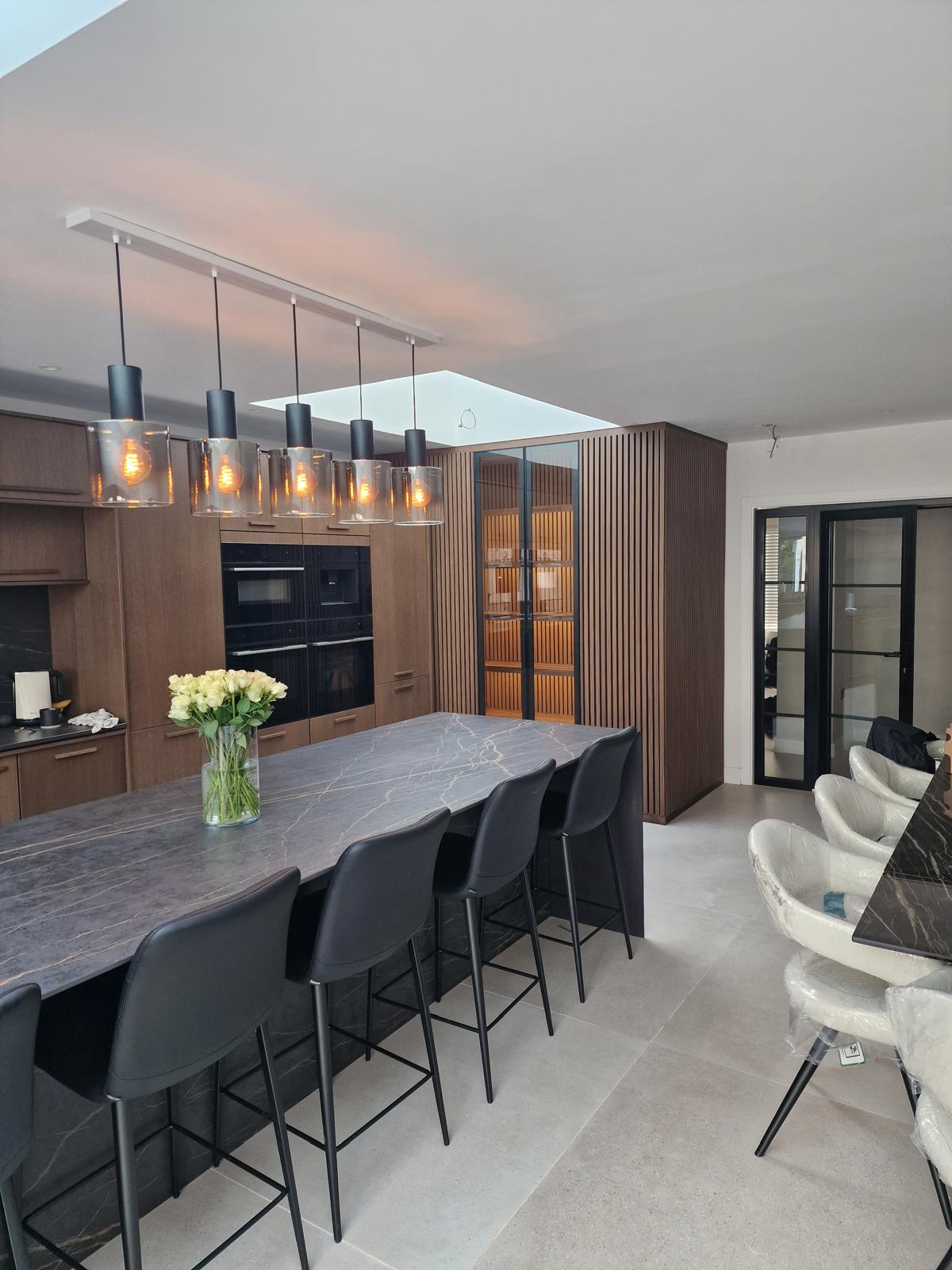 Dark walnut kitchen with tall oven housings, slatted timber panelling and a glazed pantry door beside a marble-topped island