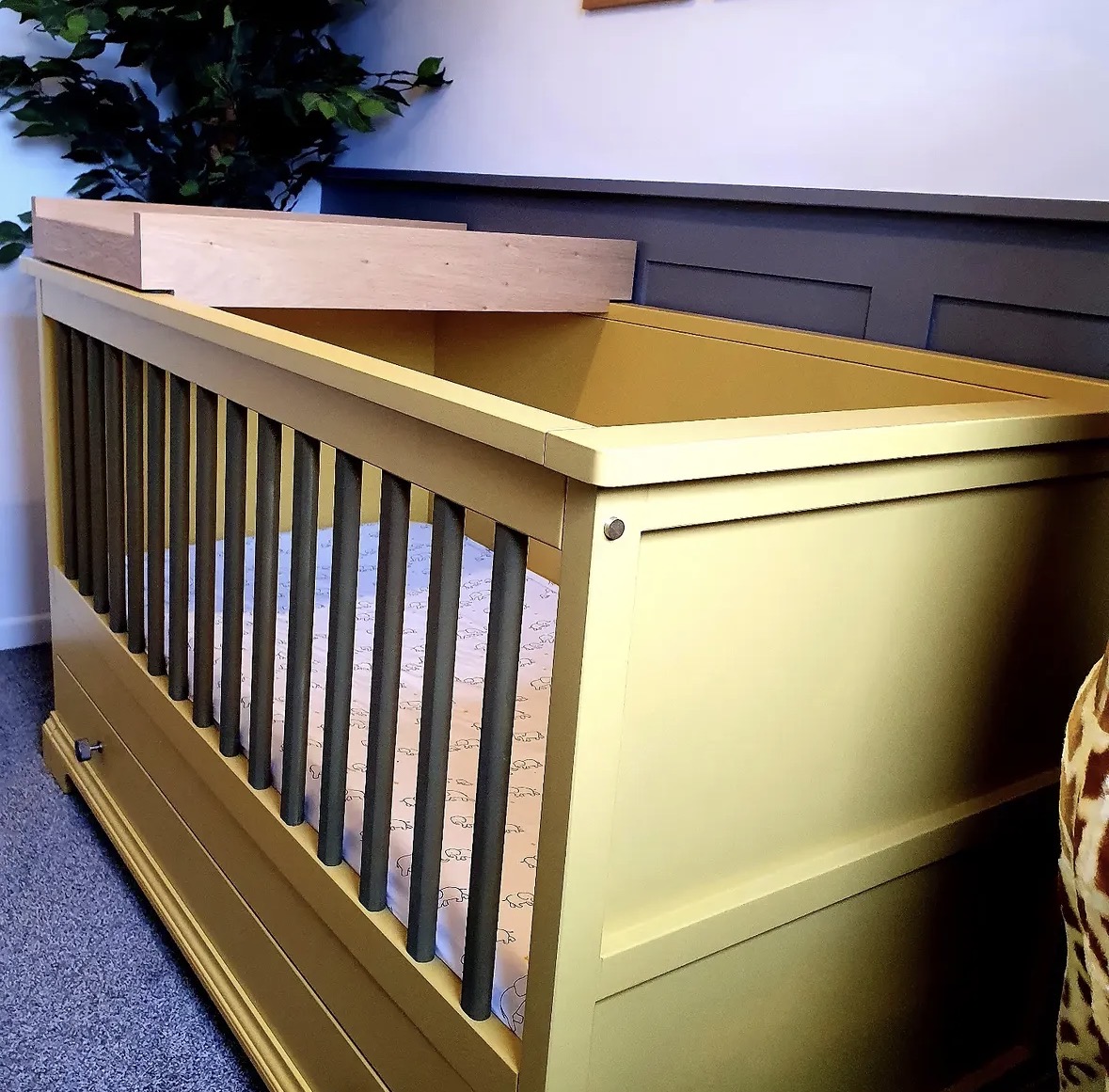 Hand-built cot bed in mustard with dark spindle rails and an oak changing top, part of bespoke nursery furniture