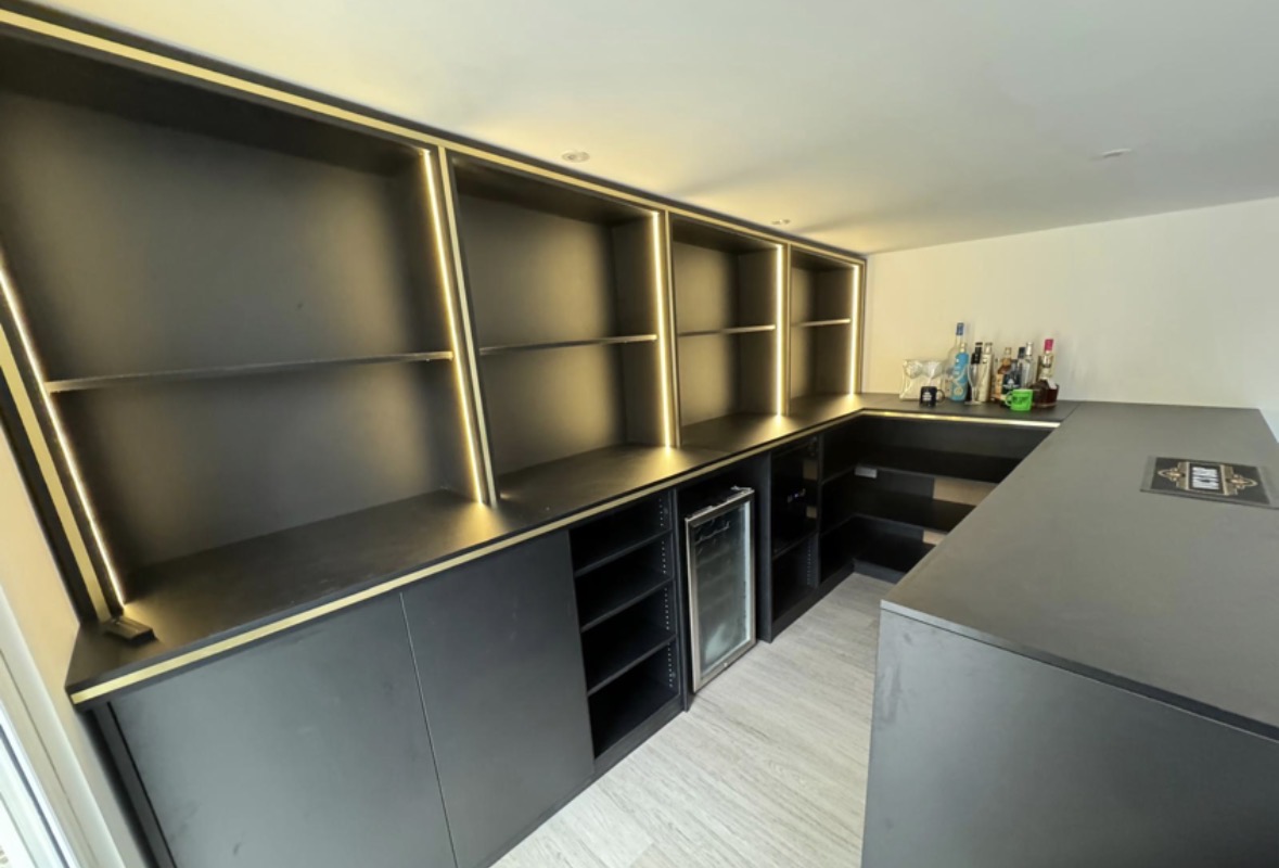 Bespoke graphite home bar with brass-trimmed open shelving lit by concealed LEDs, handleless base cabinets and an integrated glass-fronted drinks fridge