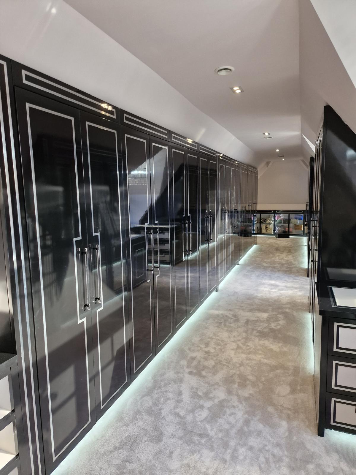 Long run of gloss black dressing room wardrobes with silver inlay, plinth lighting and a glazed display cabinet beyond
