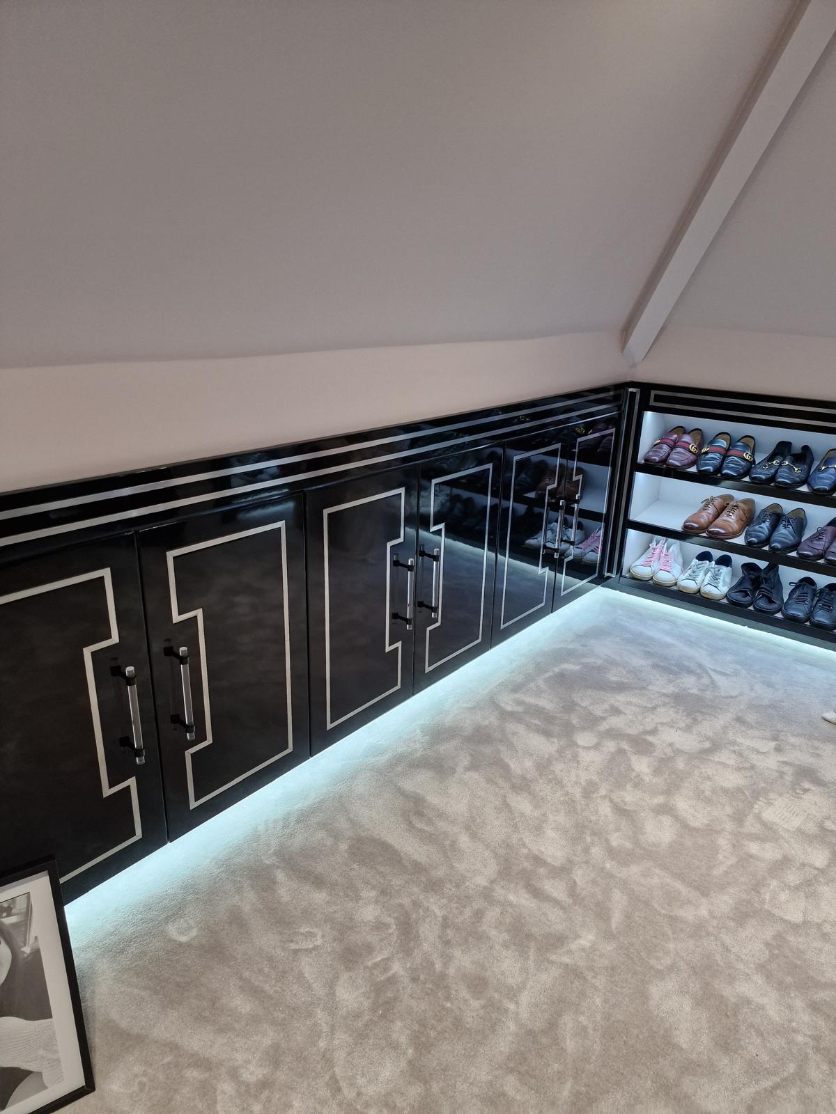 Gloss black eaves cupboards with silver inlay detailing beside LED-lit open shoe shelving in a loft dressing room