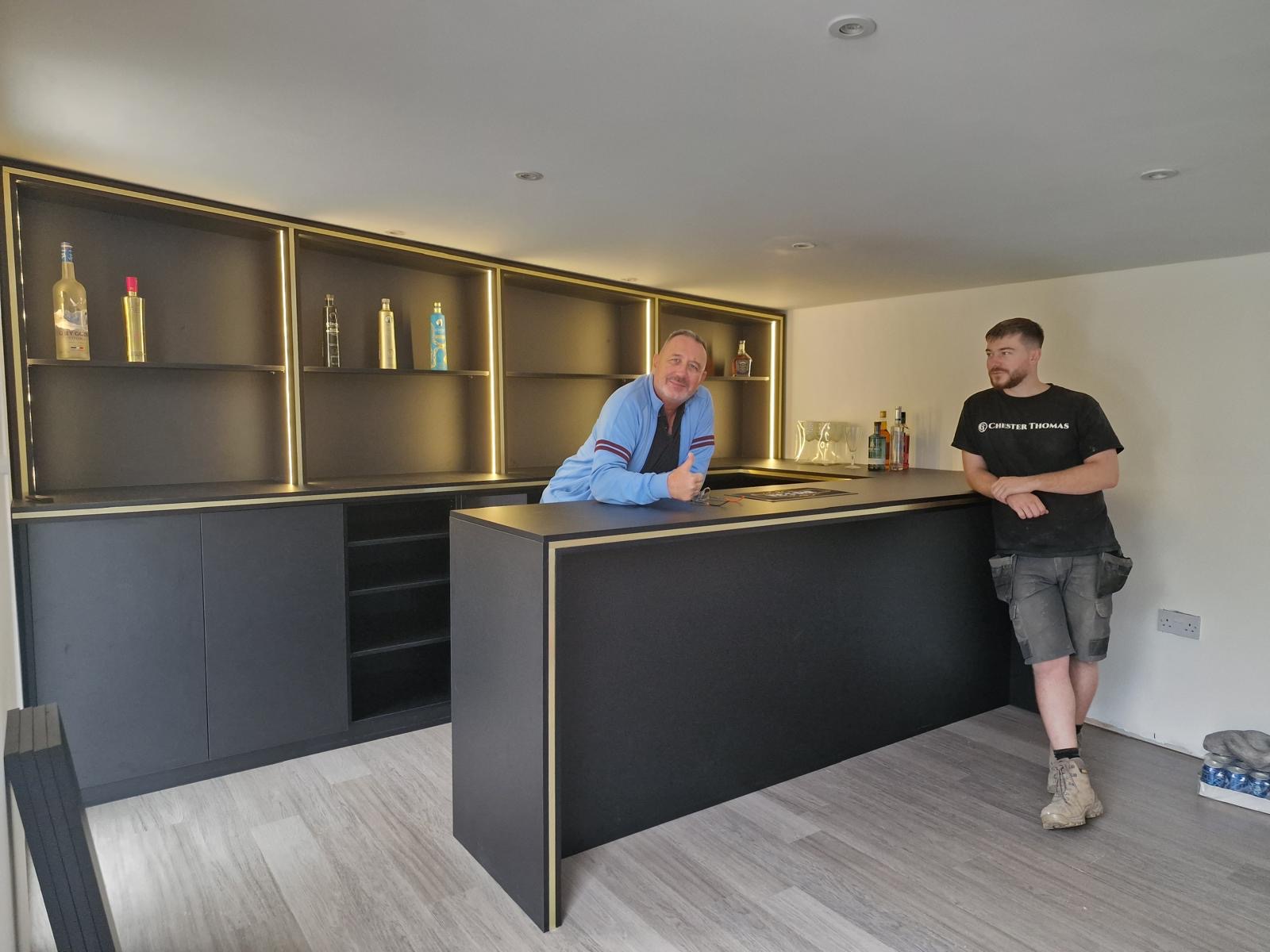 Dark fitted bar with brass trim, lit bottle shelving and a stone counter, photographed during installation