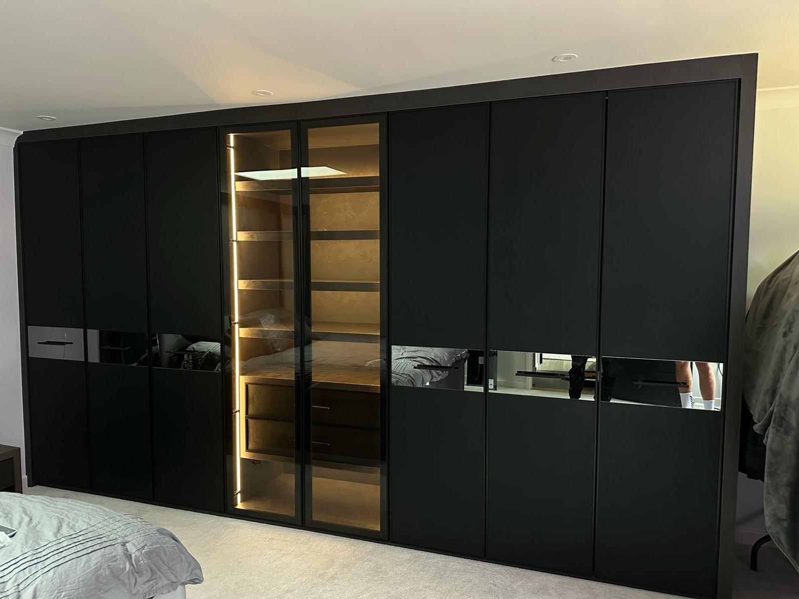 Full-height matt black wardrobe run with mirrored bands and a lit glazed centre bay showing shelving and drawers