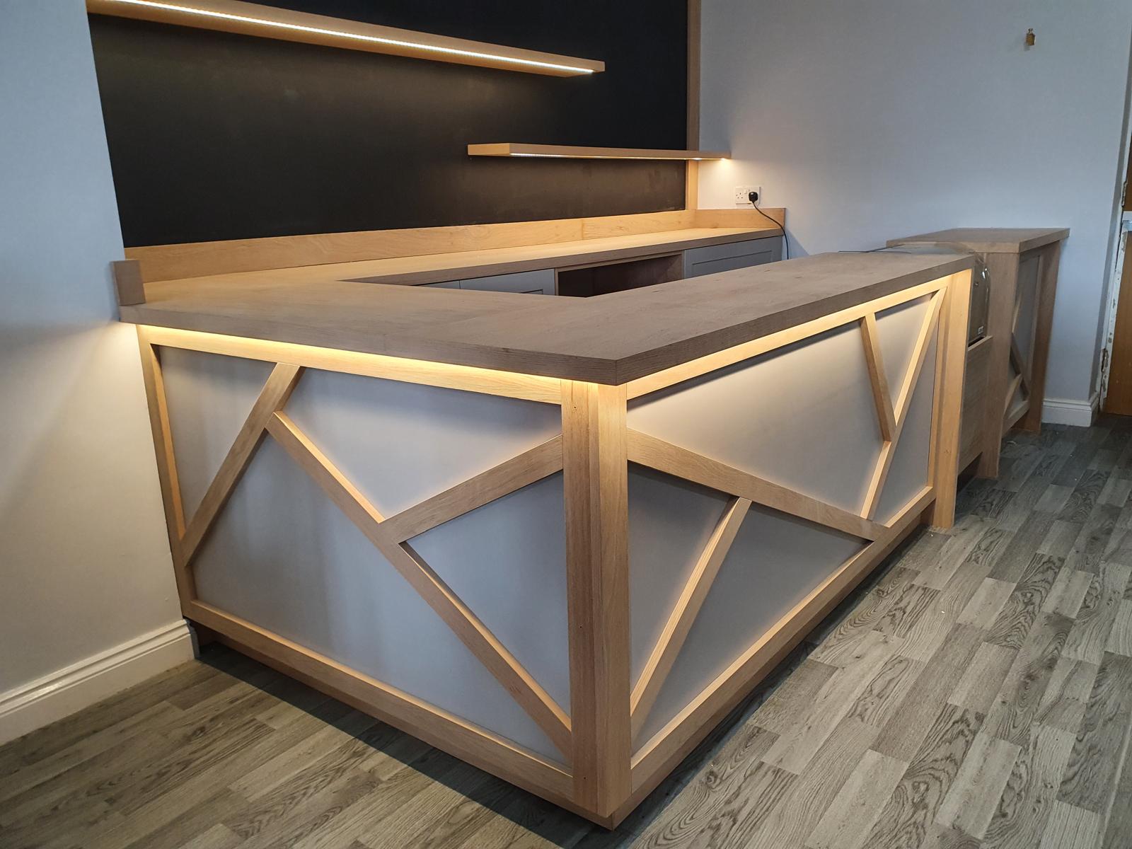 L-shaped oak bar counter with geometric framed panels, LED-lit underside and lit oak back shelving