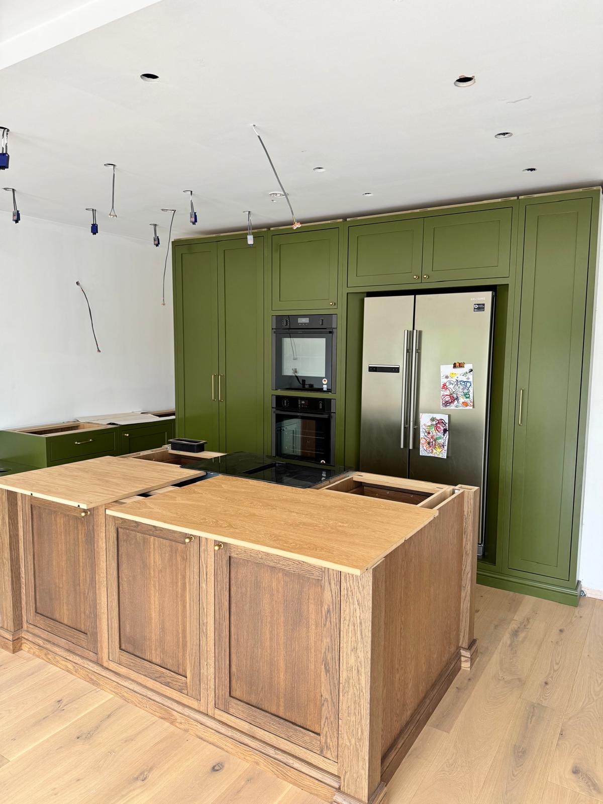 Oak island with brass fittings facing a run of olive green tall cabinets housing built-in ovens and an American-style fridge freezer