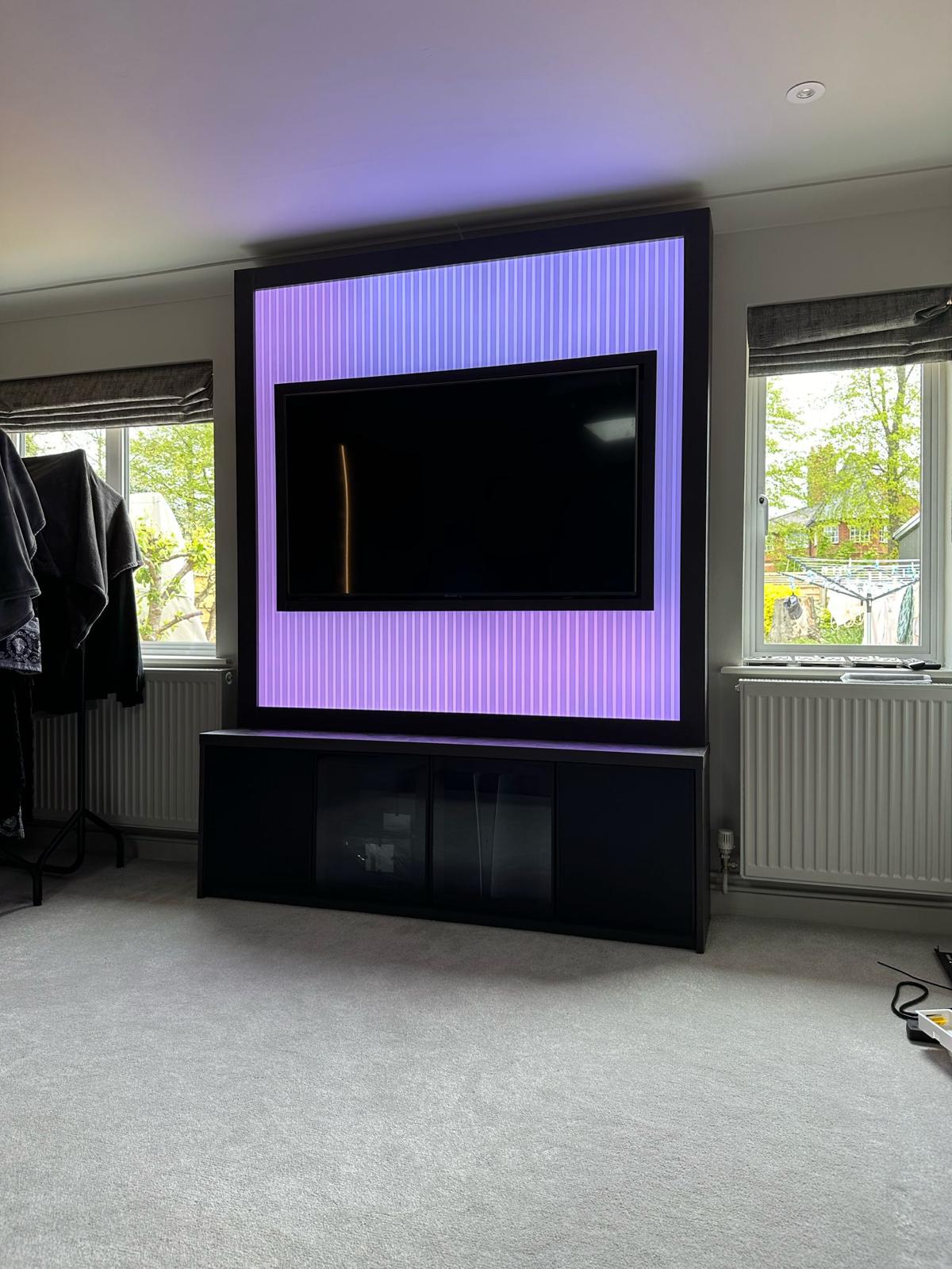 Fluted media wall panel lit lilac by concealed LEDs above a low black media unit with smoked glass doors