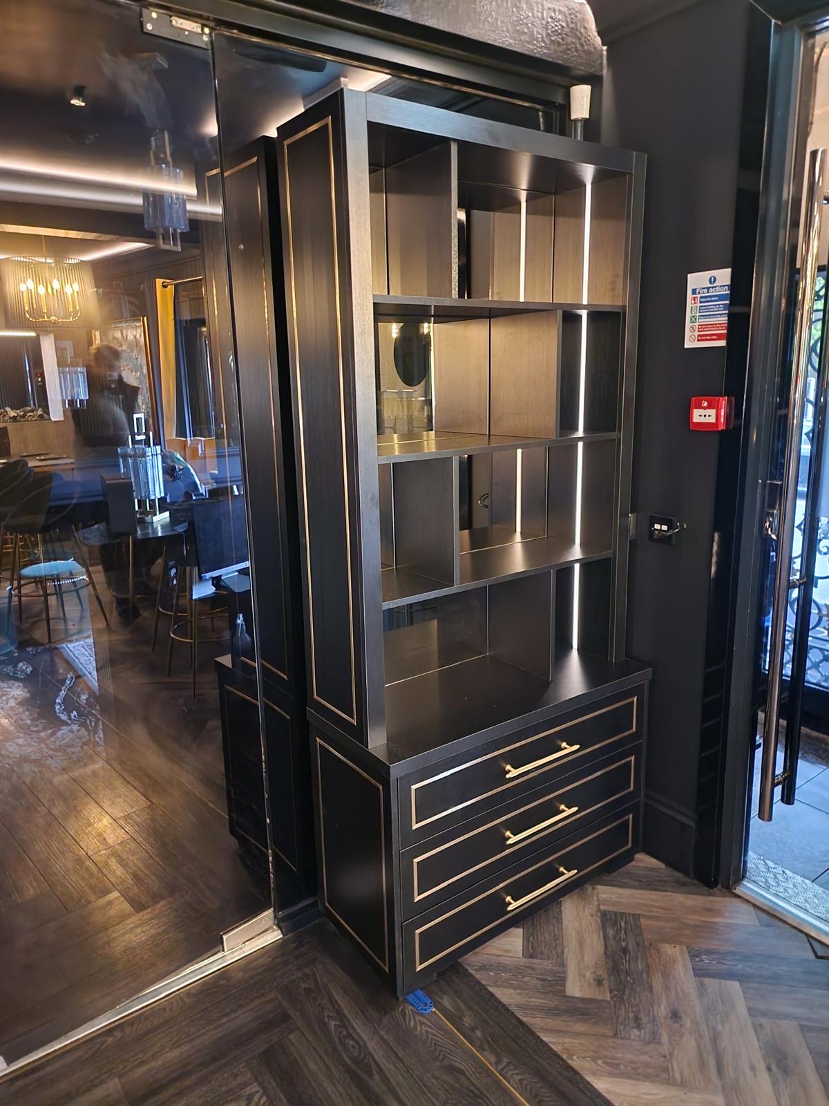 Black display cabinet with brass framing and LED-lit glass shelves holding glassware