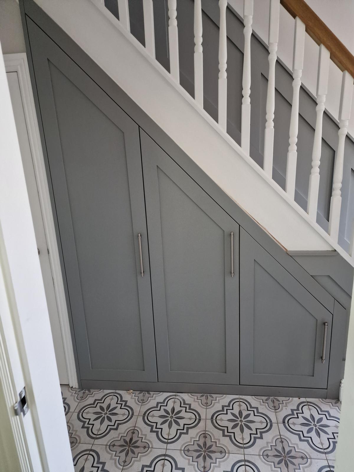 Grey painted shaker under-stairs cupboards with raked doors and brushed steel bar handles in a tiled hallway