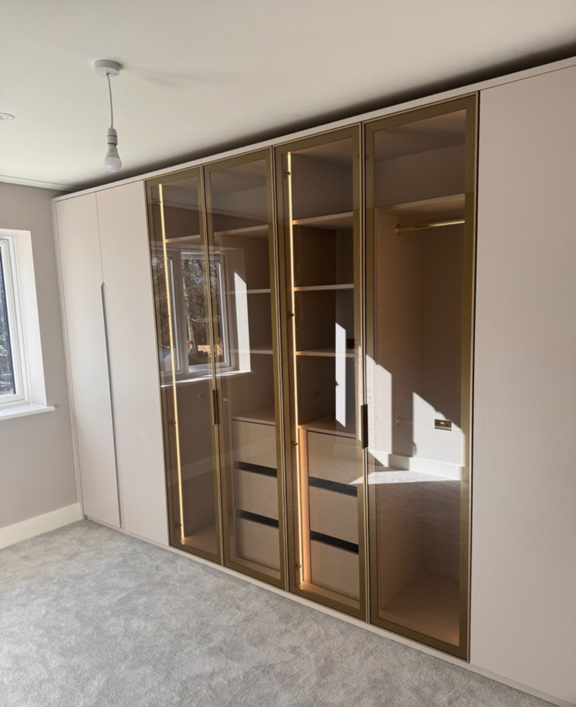 Fitted cream wardrobes with brass-framed bronze glass doors, LED-lit interiors, hanging rails and internal drawers