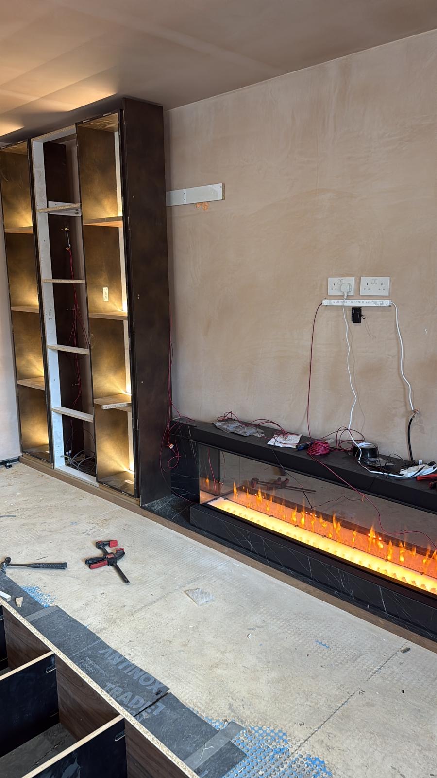 Inset linear fireplace lit in its black marble surround beside bronze-lined display shelving during installation of the Solihull media wall