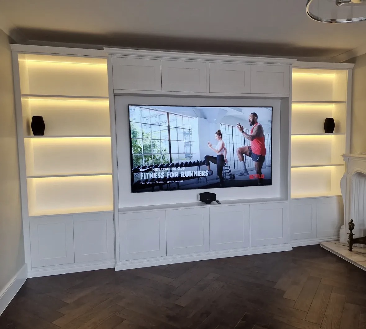 Ivory shaker-style media wall with a recessed television, warm-lit display shelves and herringbone floor