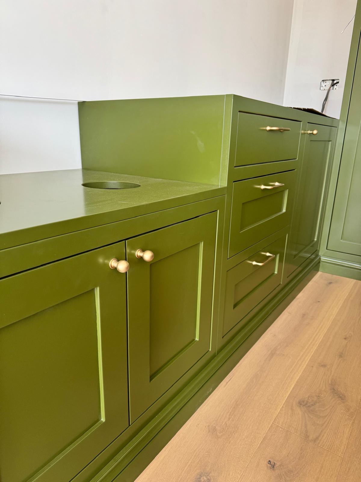 Olive green painted kitchen base cabinets and drawers with aged brass knobs and cup handles, with the sink aperture cut ready for the tap