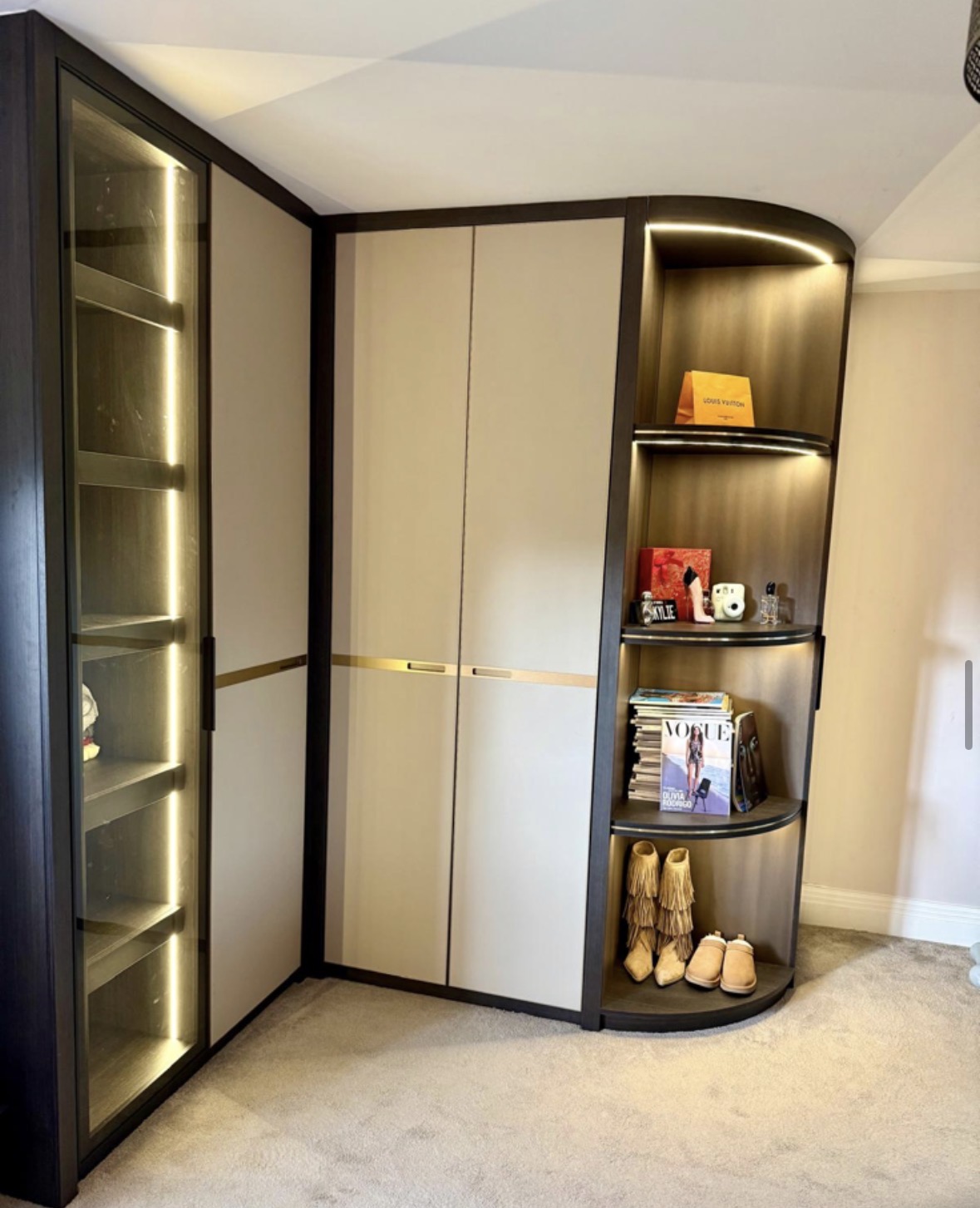 Corner run of fitted wardrobes in cream and dark oak with brass inlay handle rails, a glazed LED-lit shoe cabinet and curved open display shelving