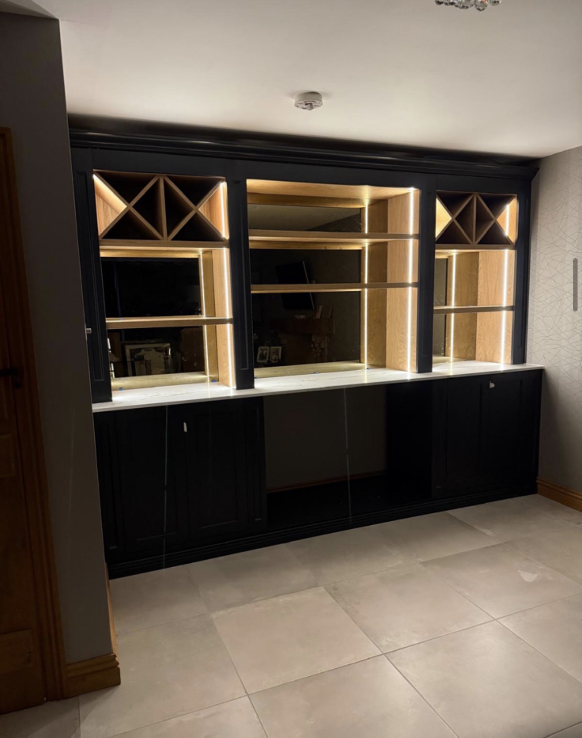 Full-wall black painted bar with oak wine racking, LED-lit mirrored shelving, marble-effect counter and panelled cupboards below