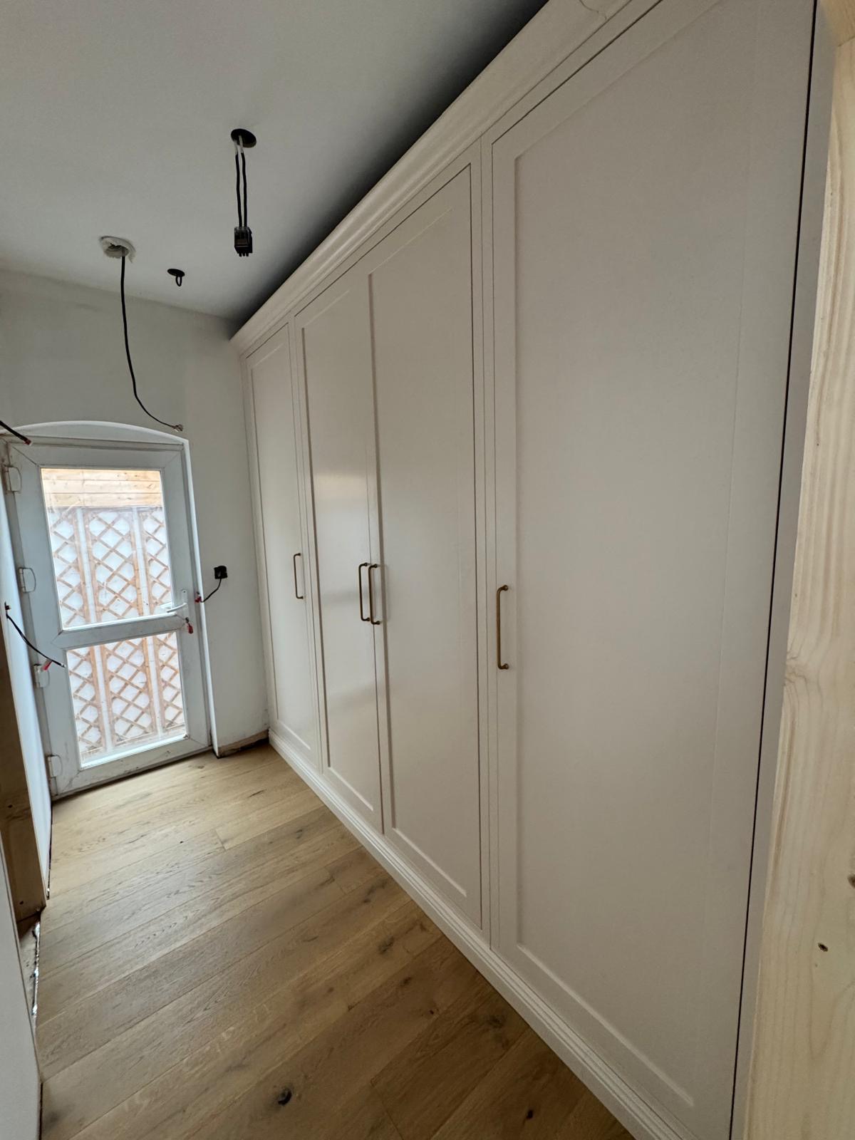 Fitted cream shaker wardrobes with brass handles running the length of a bedroom wall beside a window