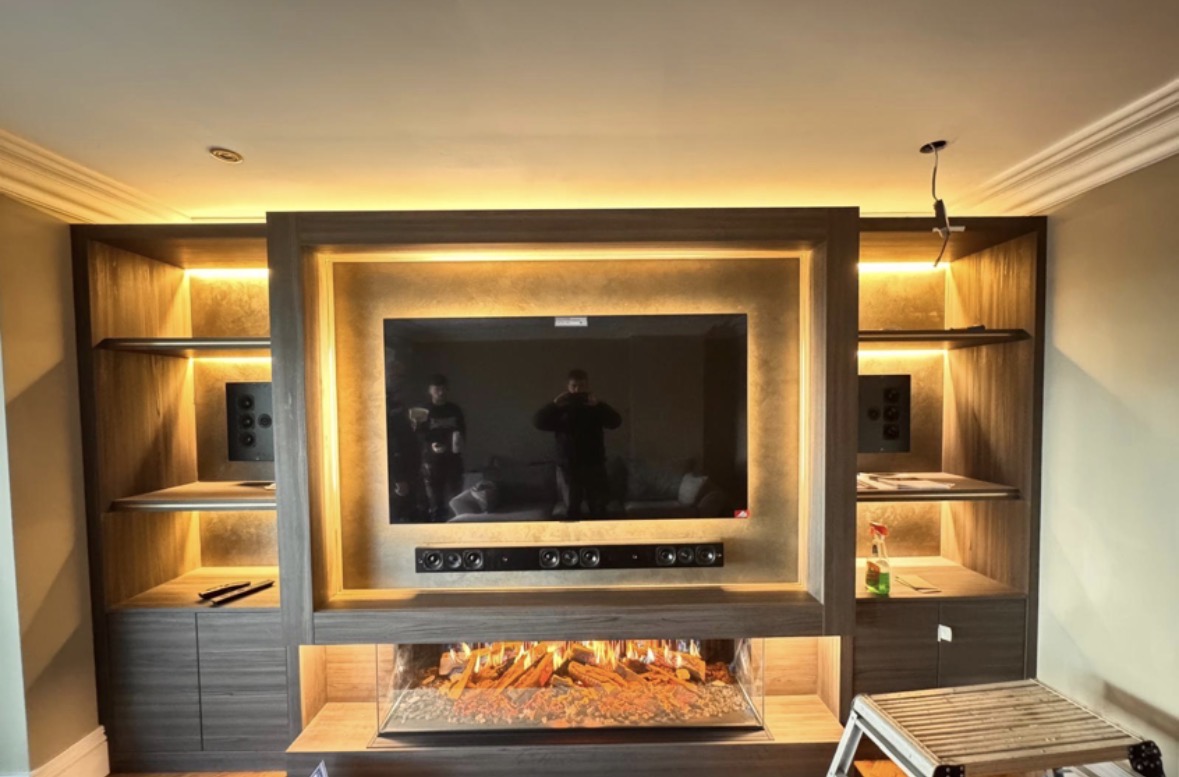 Media wall with a burr veneer television surround, long inset flame fire and LED-lit open shelving in warm timber