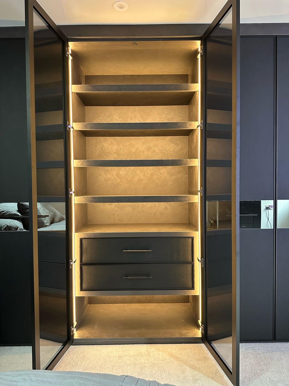 Dark glazed wardrobe doors with slim gold framing and a lit interior showing hanging rails and shelving