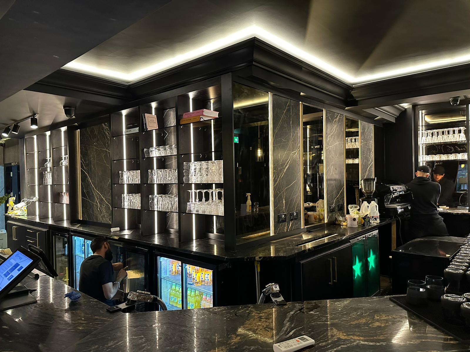 Back bar run with marble wall panels, brass detailing and LED-lit glass shelving for bottles and glassware
