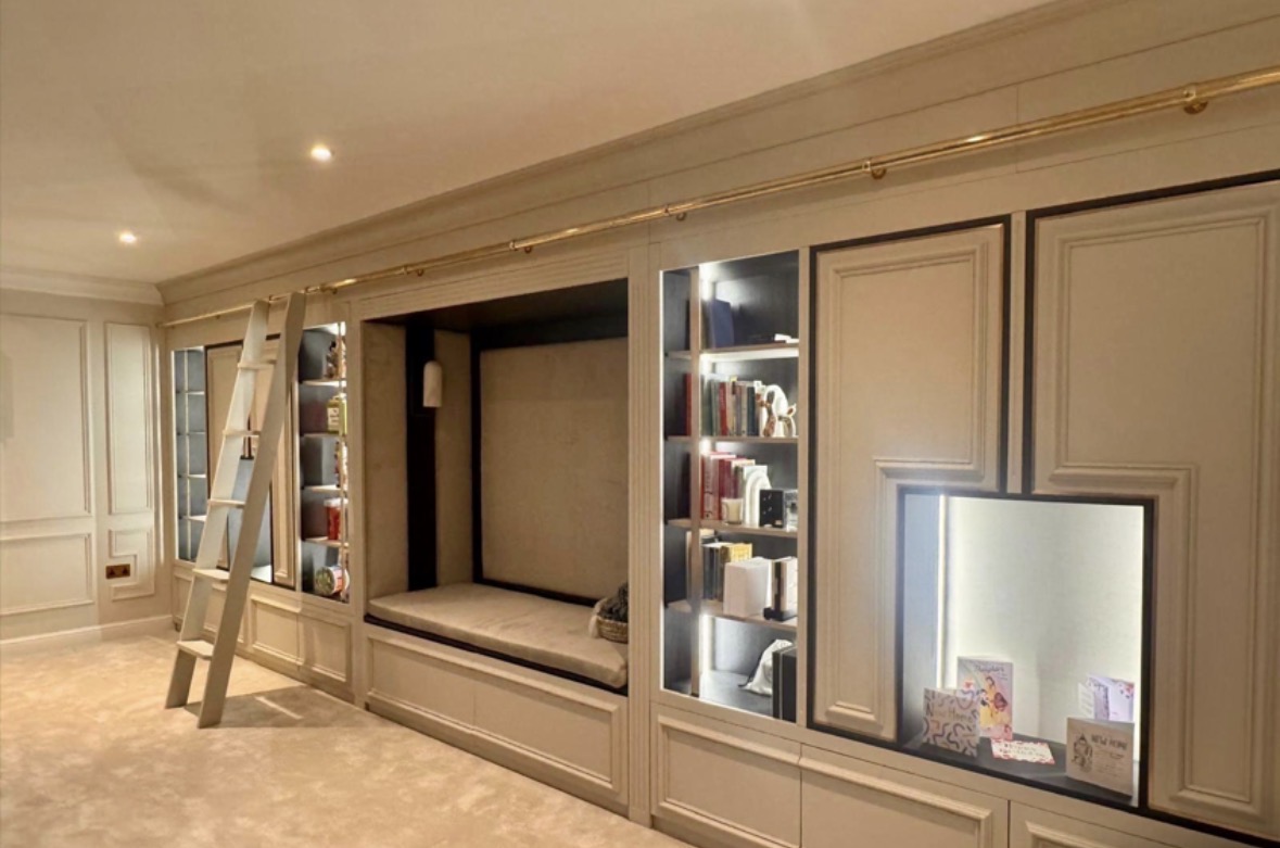 Full-wall painted study cabinetry with panelled doors, a recessed television and desk area, glazed display cabinets and a library ladder