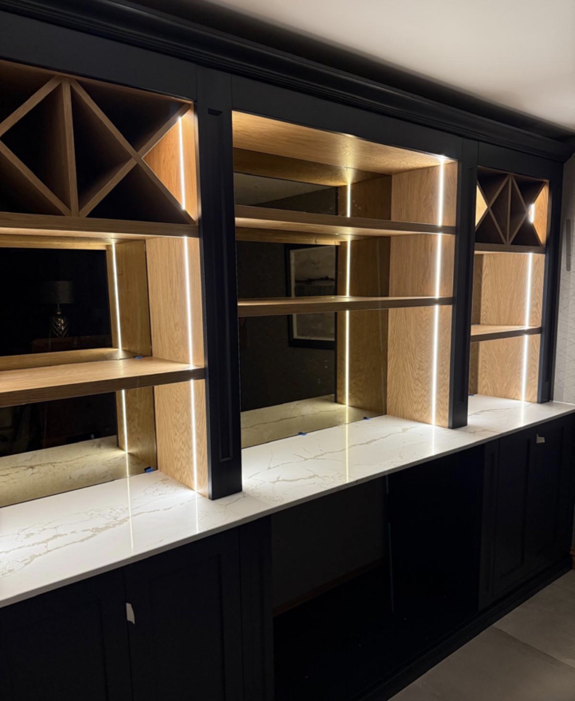 Angled view of a navy painted drinks wall with lit oak wine racking, open shelving and panelled cupboards beneath a marble-effect counter