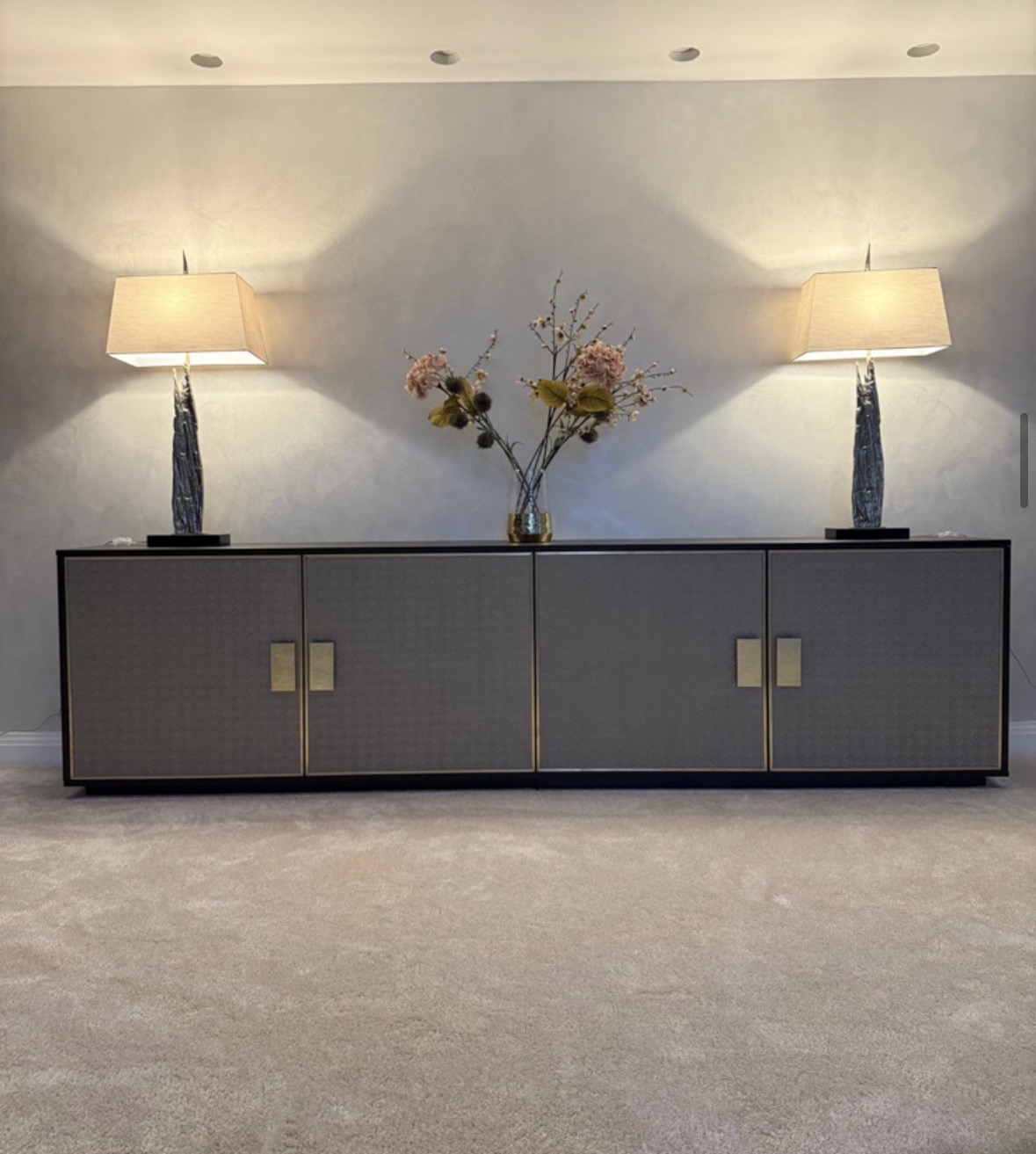 Long freestanding sideboard with woven grey textured doors, brushed brass handles and a black frame, set against a polished plaster wall