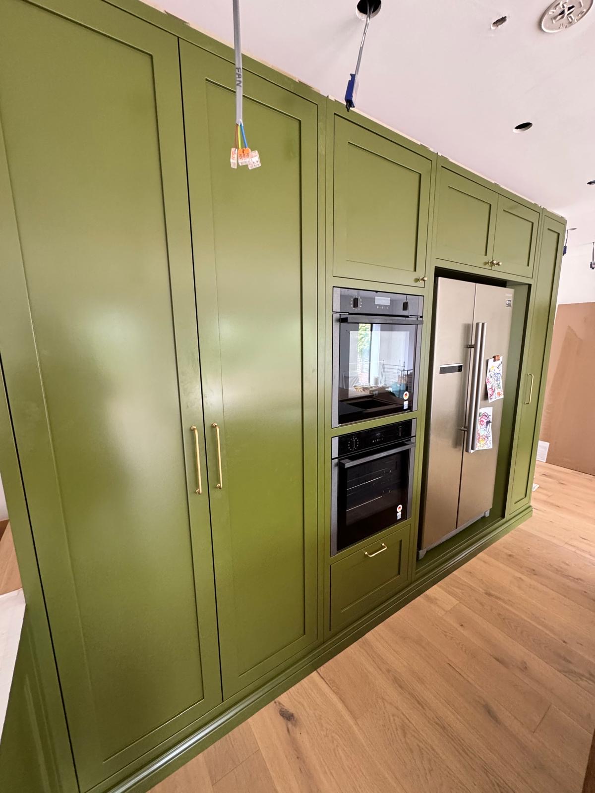 Floor-to-ceiling olive green hand-painted kitchen cabinetry with brass handles, built-in double ovens and an integrated American-style fridge freezer