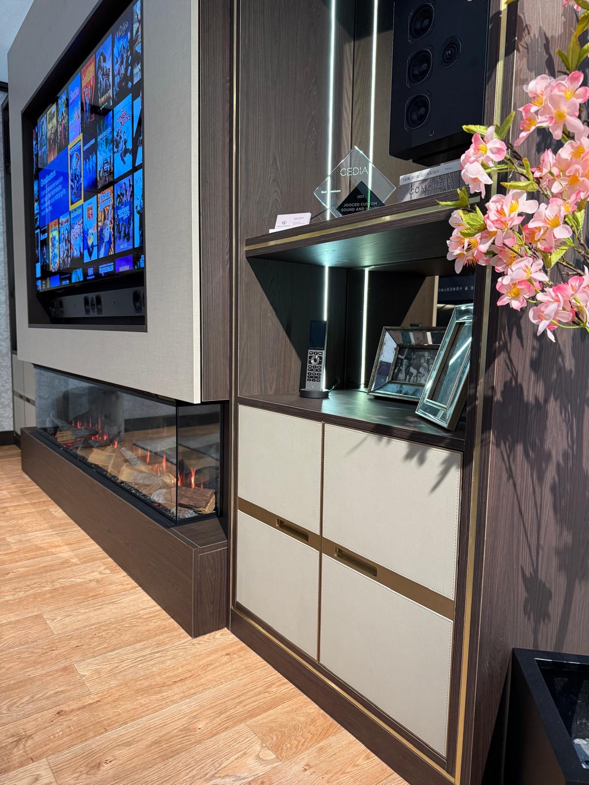 Angled view of a bespoke media wall showing the inset electric fire, dark oak carcass and LED-lit display shelving with brass detailing