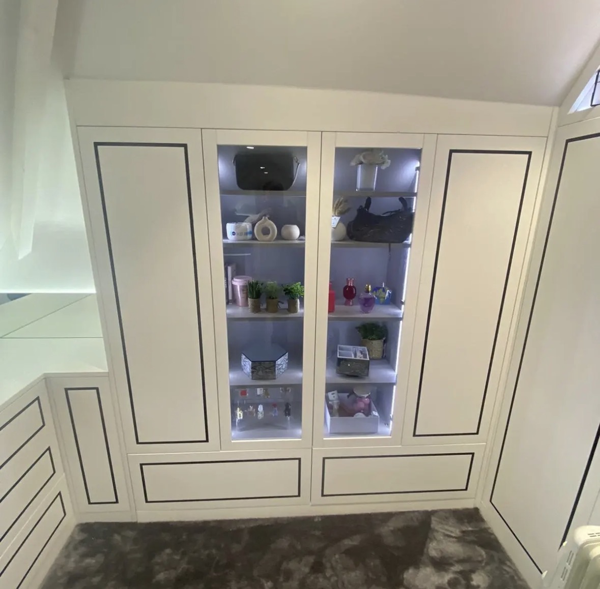 Glazed, internally lit display cabinets between fitted ivory wardrobes in a bespoke loft dressing room