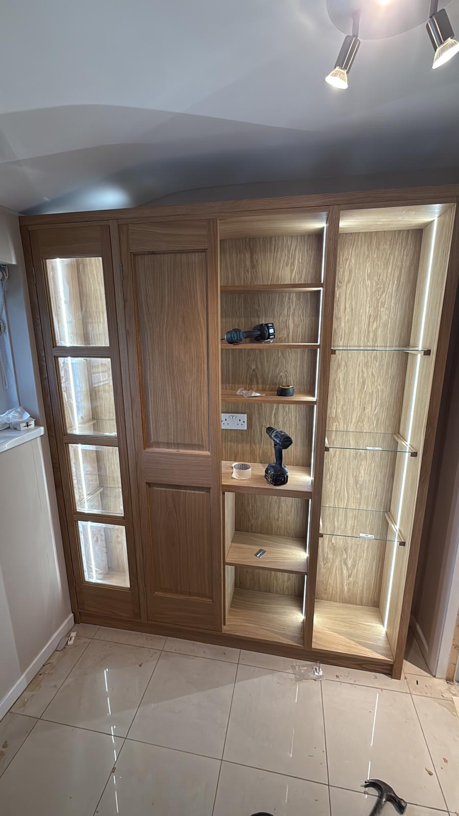 Oak display unit part-way through fitting, with one glazed door hung, LED-lit open shelving and glass shelves being installed