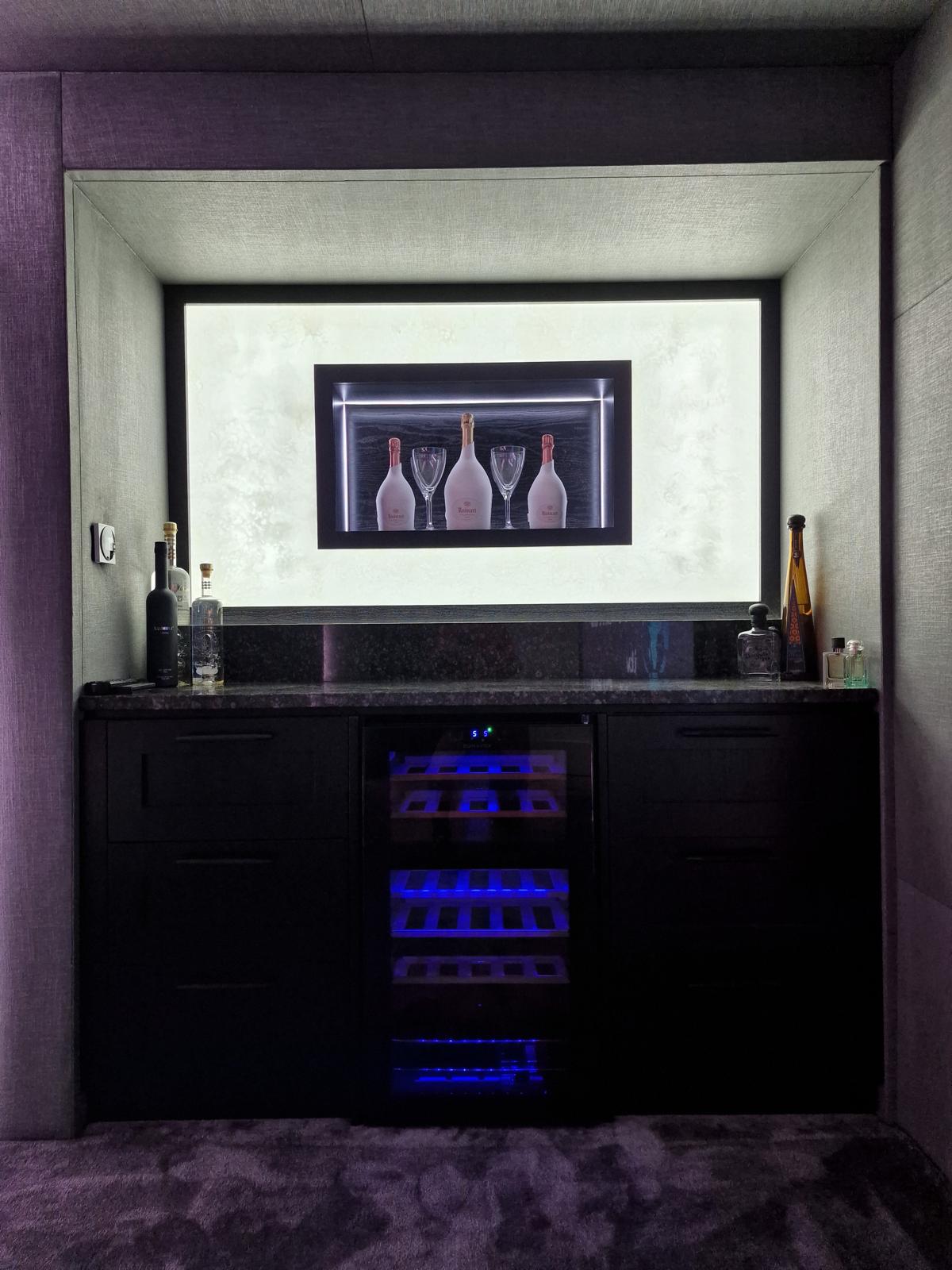 Dark drinks bar in a cinema room with stone worktop, integrated wine fridge and a lit recessed display niche above